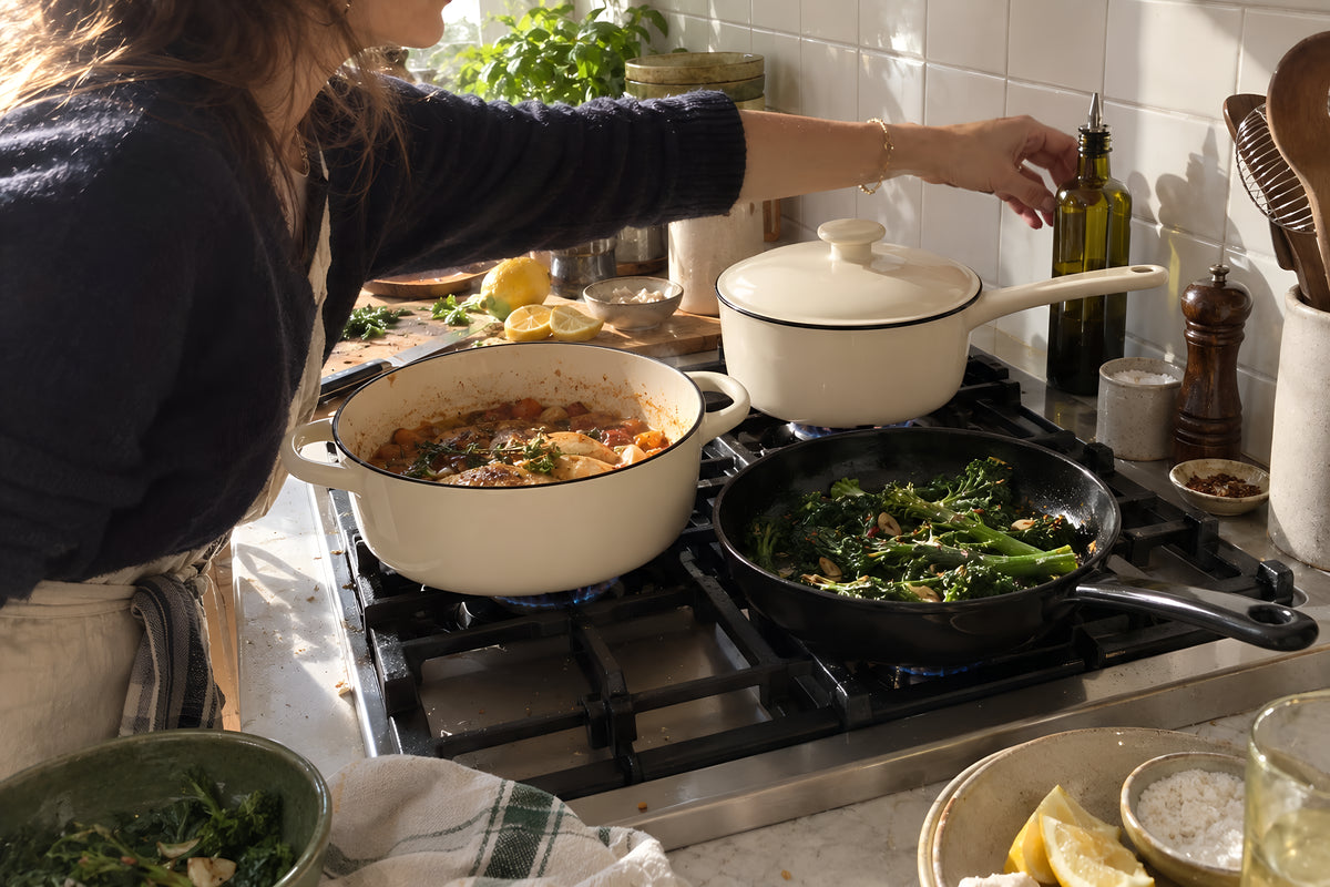 Three KILN pans going at once on a domestic hob, the cream Casserole with its lid off, the cream Saucepan and the black Frying Pan with greens in it, a woman reaching across to the far one, tiled splashback behind. Daytime, hard sun, no candles lit.