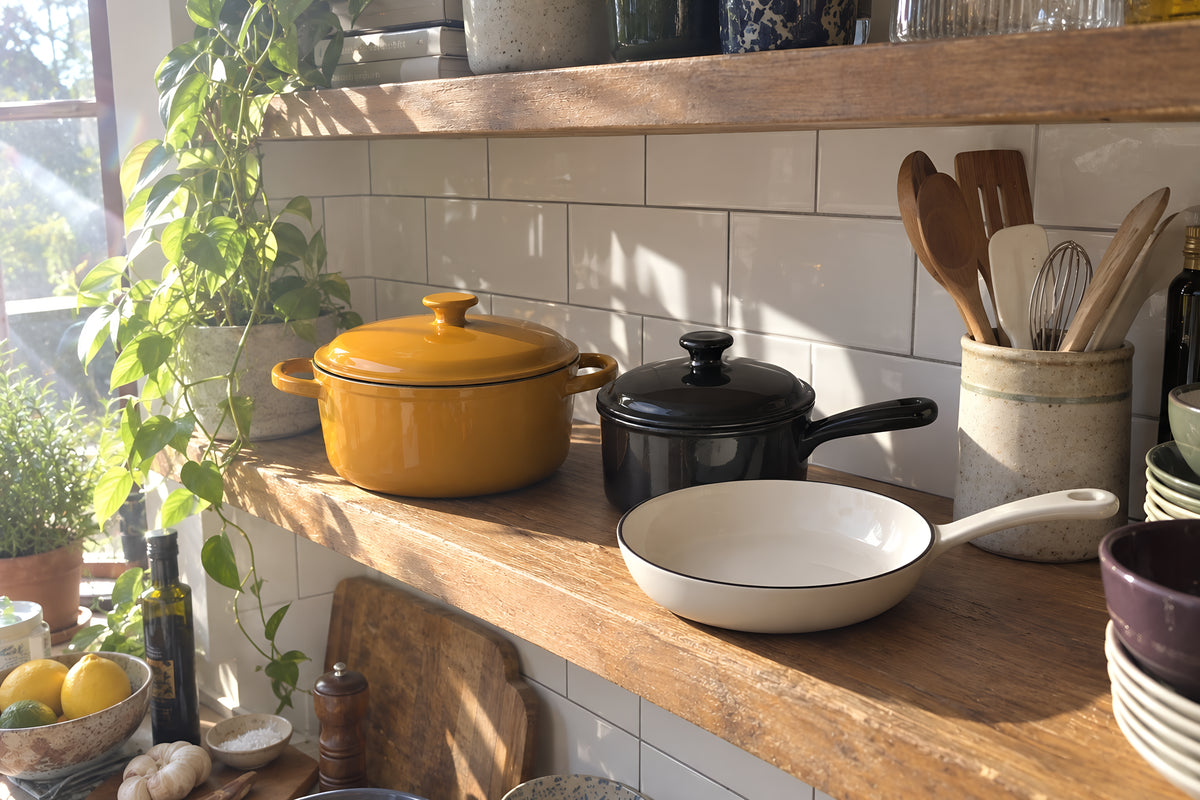 The KILN Stovetop Set out on an open kitchen shelf, mustard casserole, black saucepan and cream frying pan, a jar of utensils beside them and a tiled wall behind. Daytime, hard sun, no candles lit.