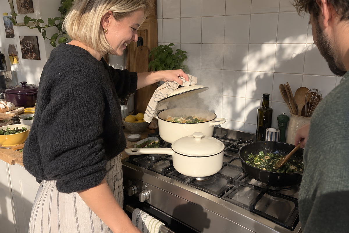 Three KILN pans going at once on a domestic hob with two people working round each other, one lifting a lid, steam and a tiled splashback behind. Daytime, hard sun, no candles lit.