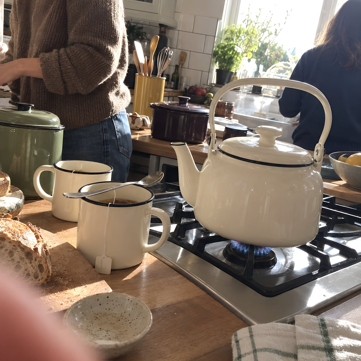 The cream KILN Kettle just boiled on a gas hob with two KILN Mugs waiting beside it, tea bags in, a spoon across one. Daytime, hard sun, no candles lit.