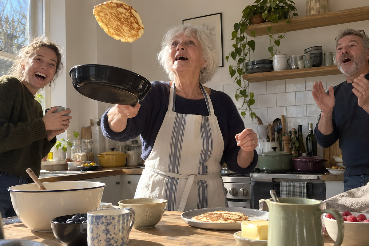 A pancake mid-air over the black KILN Frying Pan, the cook's face tipped up watching it, two others in the background braced to cheer or jeer. Daytime, hard sun, no candles lit.