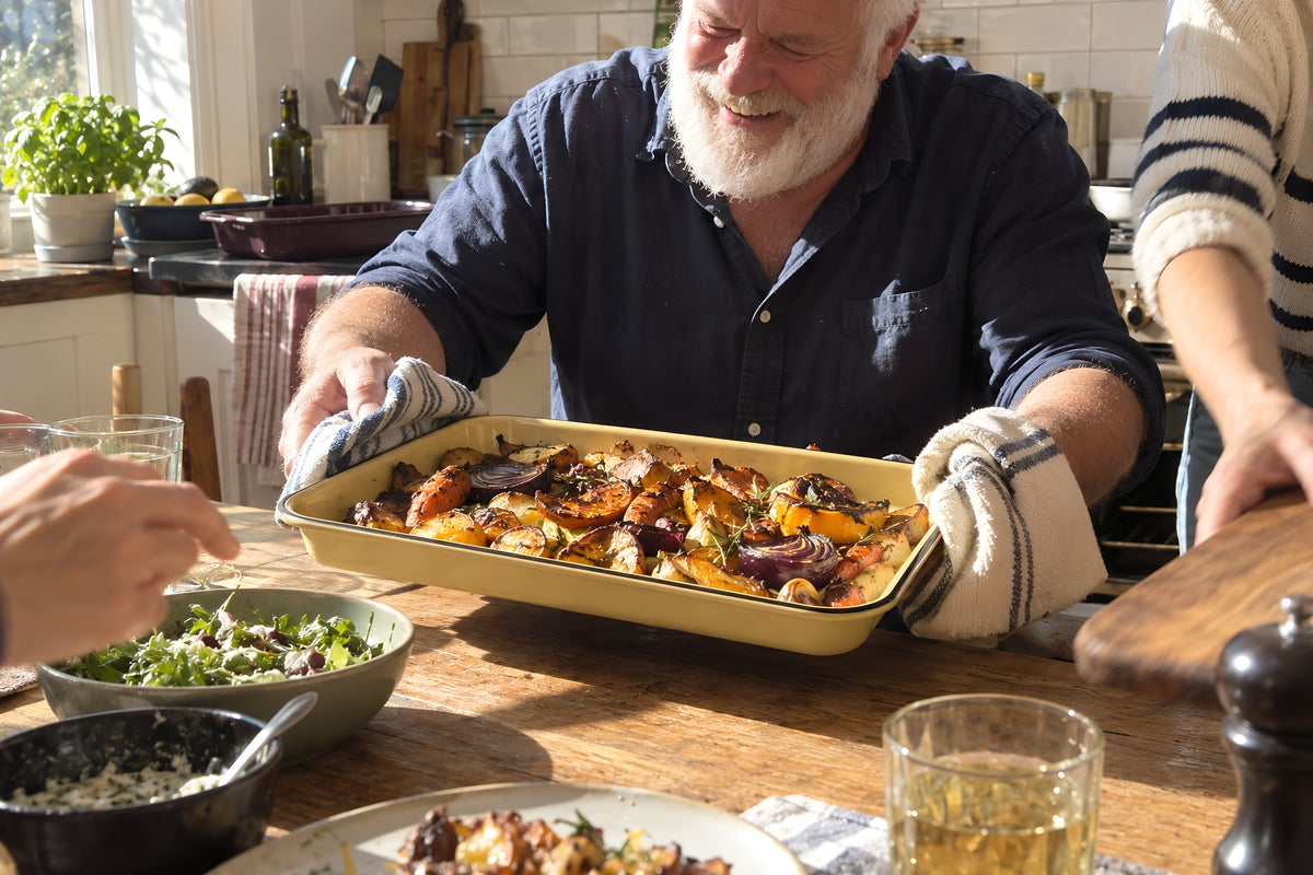 A tray of roast vegetables coming out of the oven on the mustard KILN Tray, oven gloves on, another pair of hands moving a board out of the way. Daytime, hard sun, no candles lit.