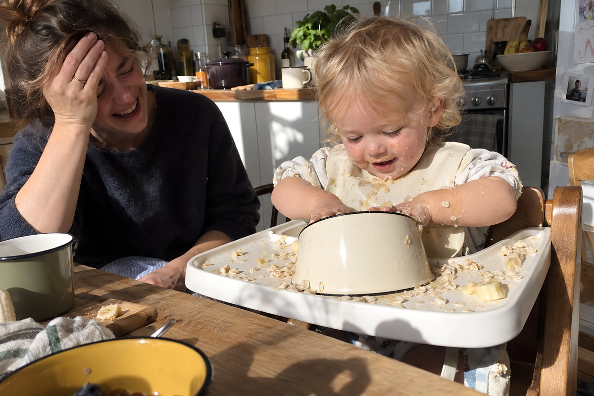 A toddler in a high chair with a cream KILN Bowl upended on the tray, hands in it, a parent halfway between laughing and despairing. Daytime, hard sun, no candles lit.